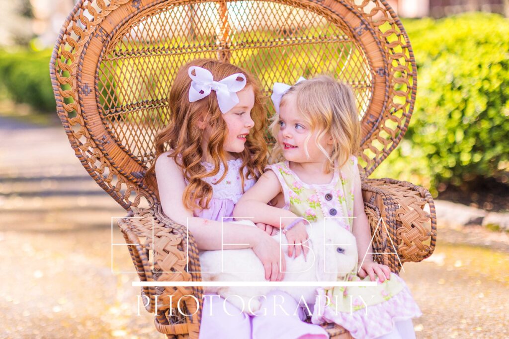 Sweet sisters smile at each other while holding a white bunny and sitting in a brown wicker fan back chair at live bunny mini sessions in Chickamauga, Georgia