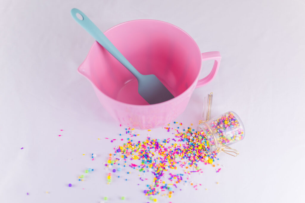Pink mixing bowl with teal mixing spoon and spilled rainbow sprinkles in glass and gold jar