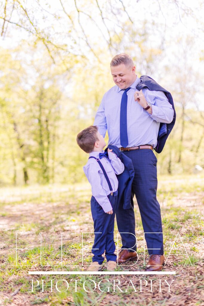 Dad and son smile at each other with navy blue suit jackets tossed over their shoulders
